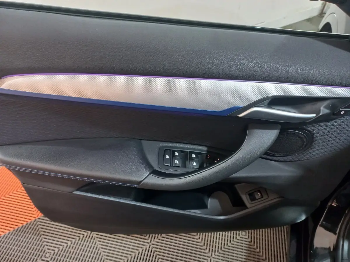 Vue intérieure côté gauche de la porte noire du BMW X1 xDrive20d M Sport avec éclairage d'ambiance bleu et commandes fenêtres.
