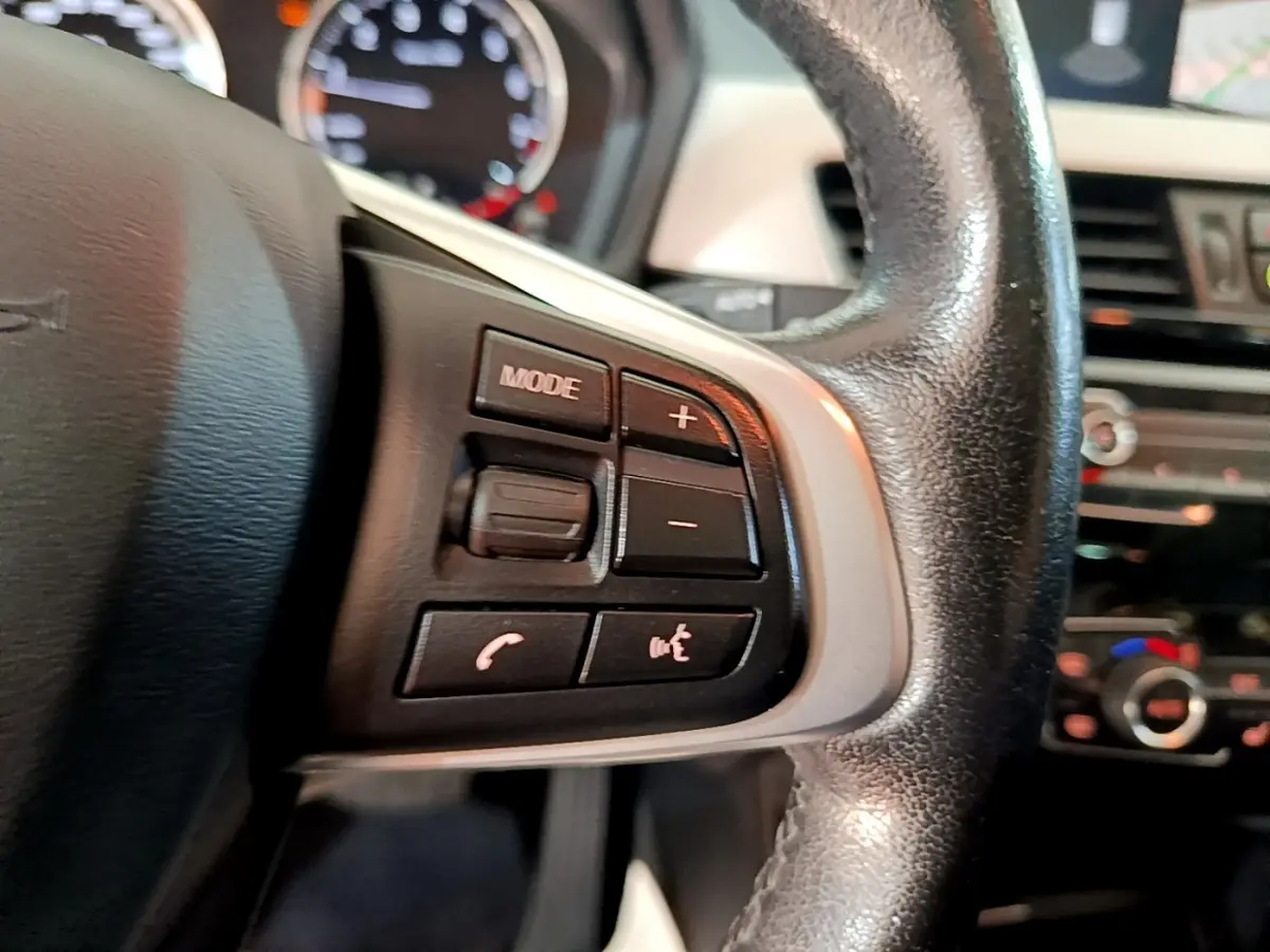 Gros plan sur les commandes multimédias au volant cuir noir d'une BMW X1 sDrive20i 2020, intérieur visible flou.