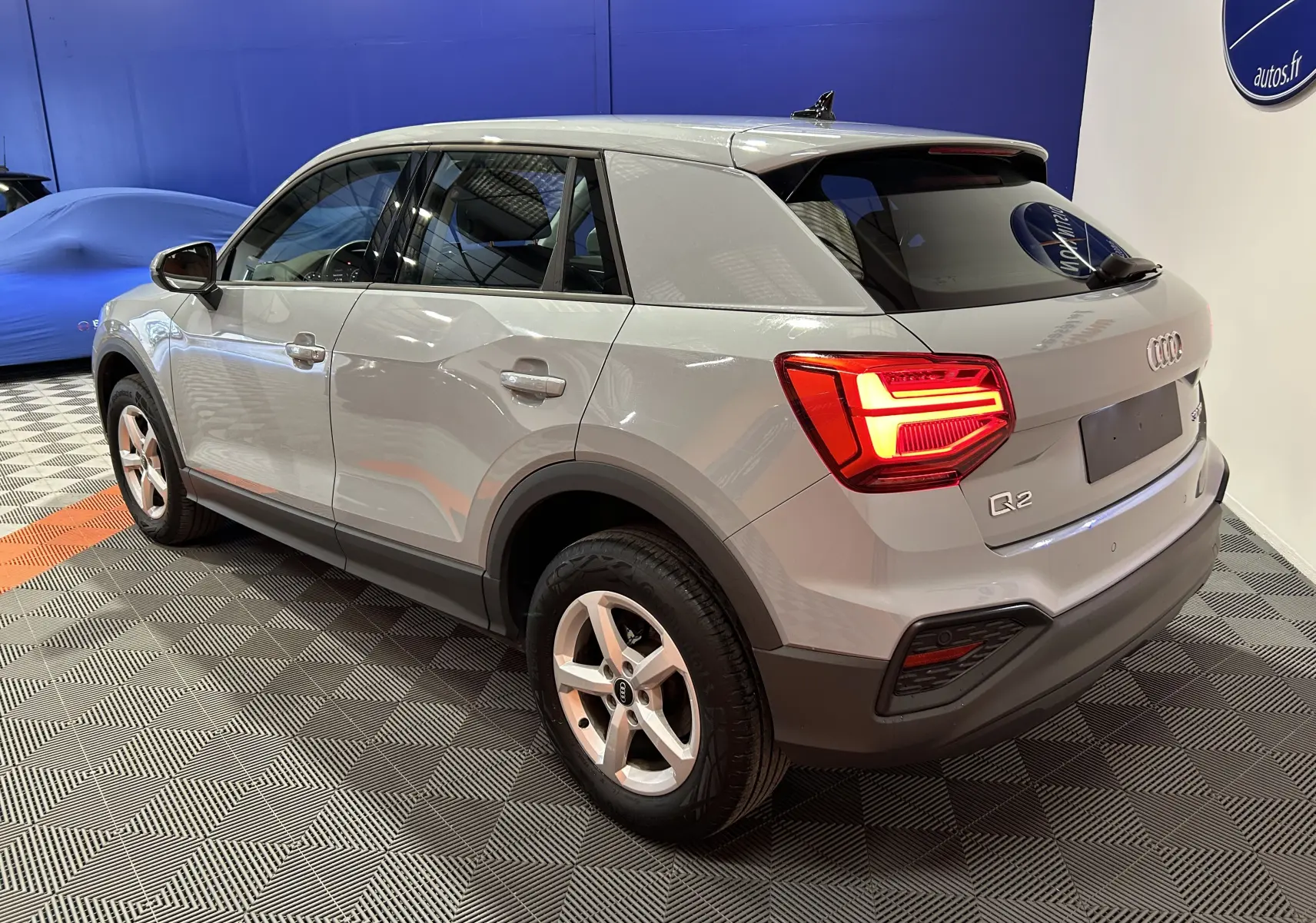 Audi Q2 gris en 3/4 arrière droit, feux arrière allumés, hayon électrique visible dans un showroom intérieur.