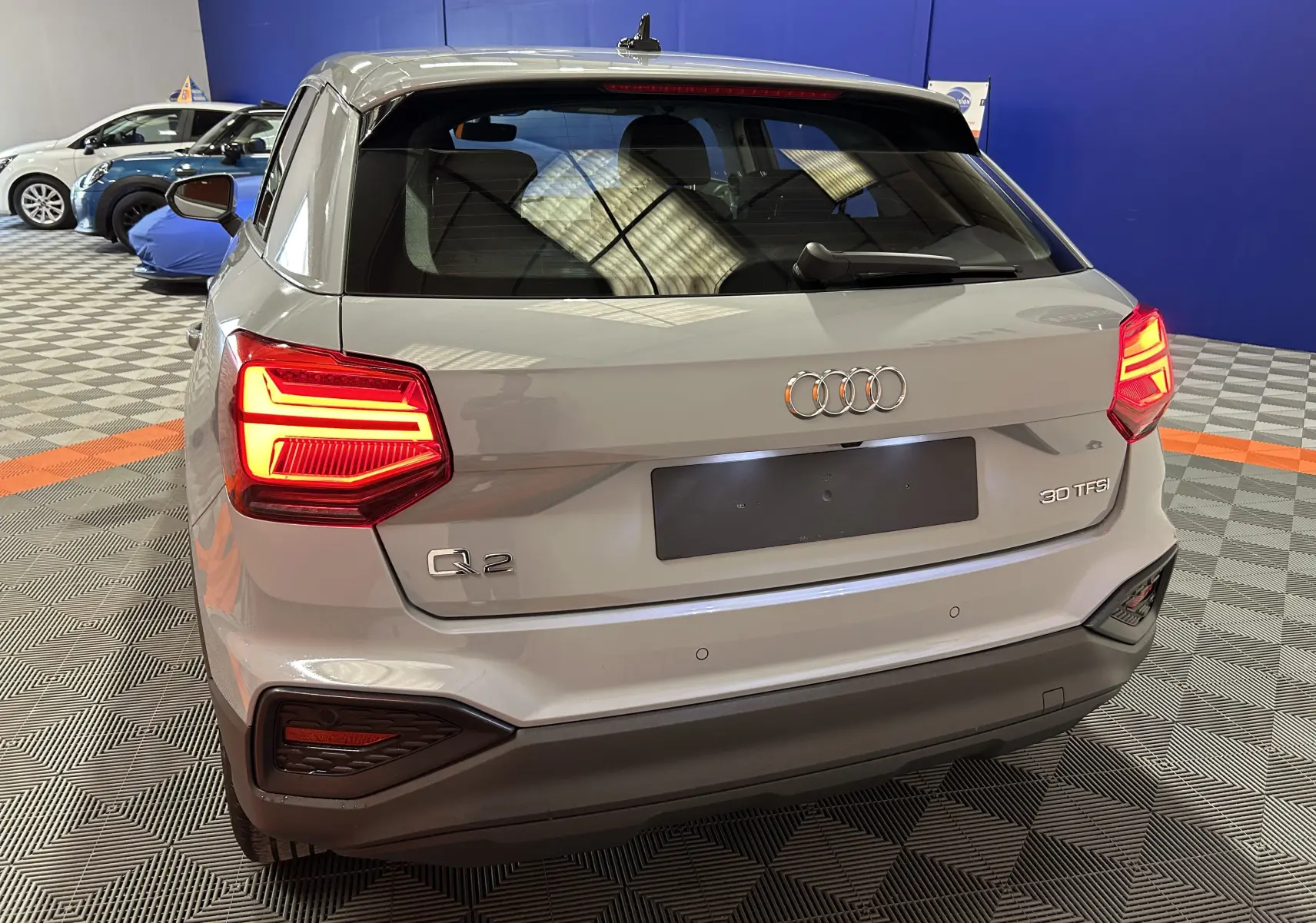 Vue 3/4 arrière d'un Audi Q2 gris avec feux arrière allumés dans un showroom intérieur.