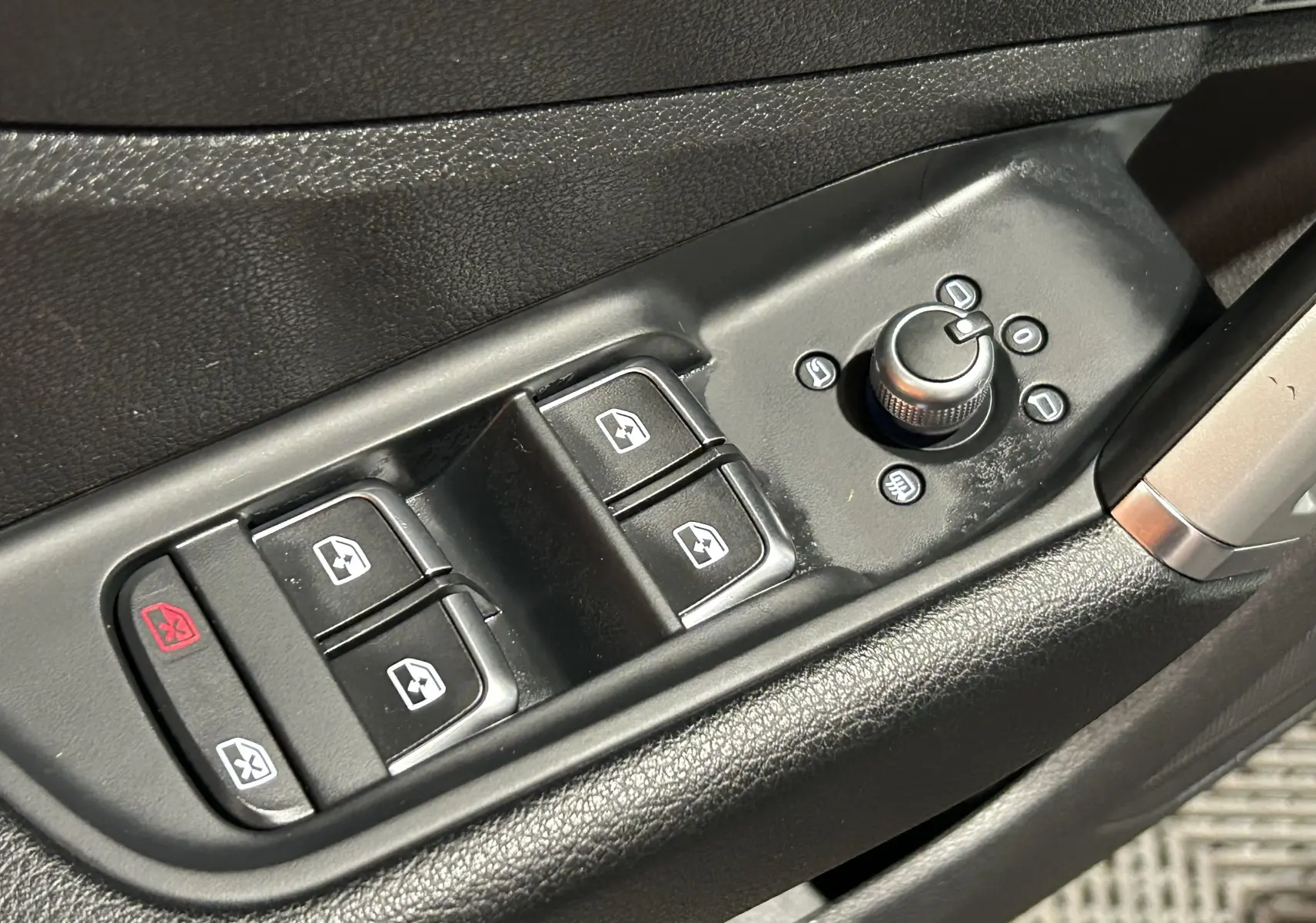 Gros plan sur les commandes de lève-vitres et réglage des rétroviseurs côté conducteur d’un Audi Q2 gris, intérieur noir.