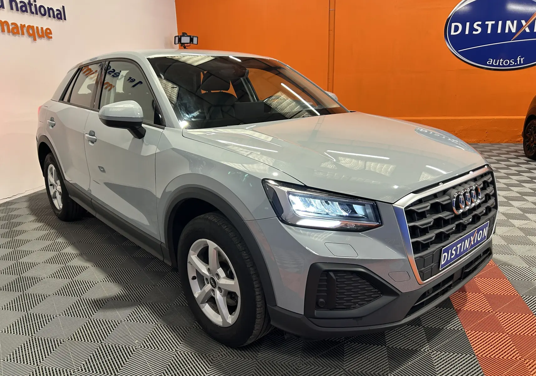 Audi Q2 gris en 3/4 avant droit, phares allumés et calandre hexagonale distinctive, dans un showroom intérieur.