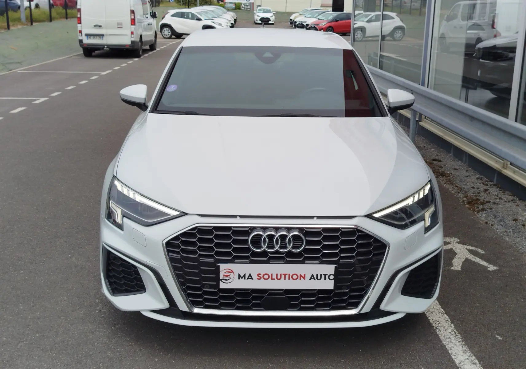 Vue frontale d'une Audi A3 Sportback blanche Glacier métallisé avec calandre hexagonale et phares LED allumés.