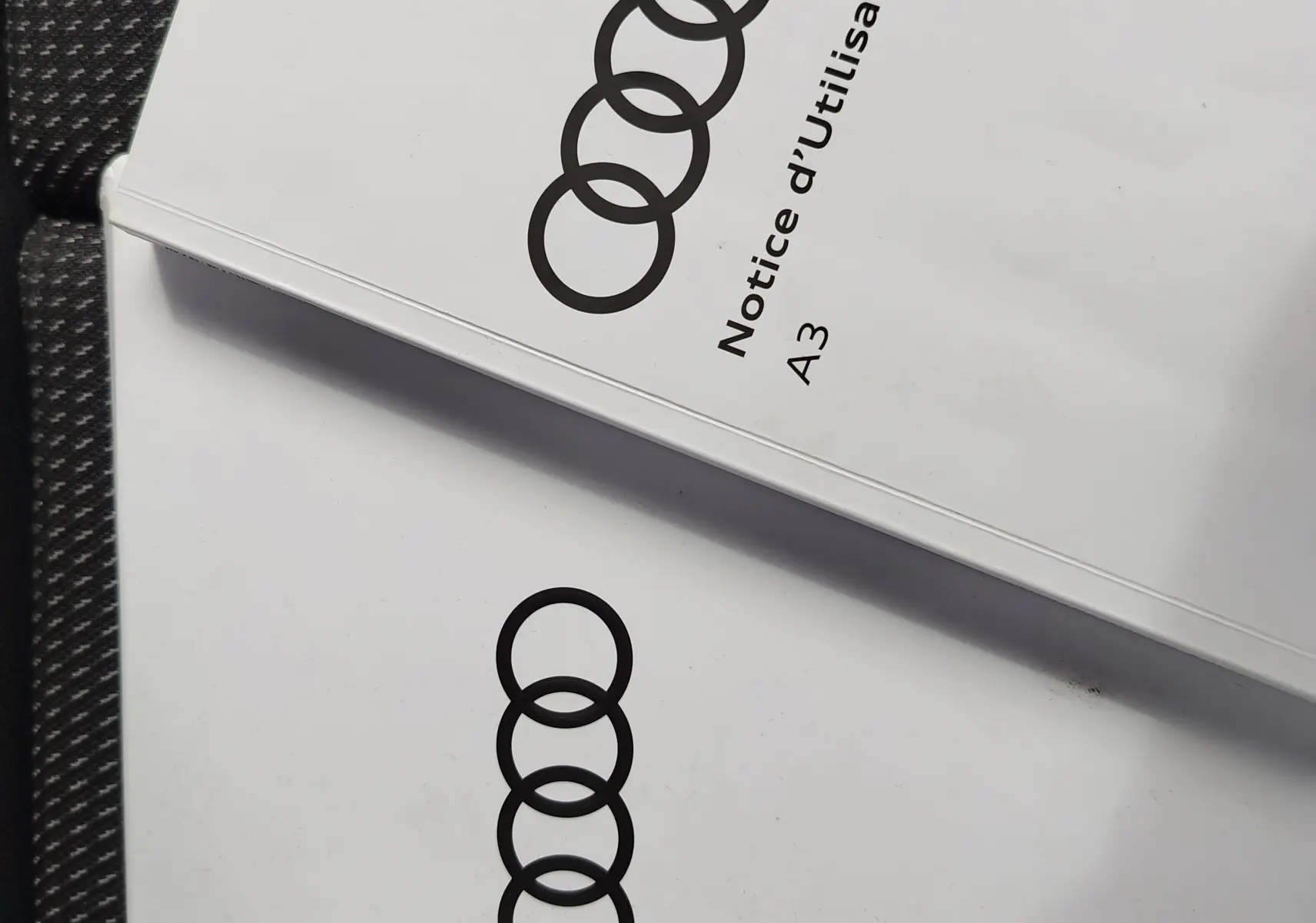 Image ne montrant pas le véhicule mais deux notices d'utilisation Audi A3 posées sur un siège en tissu gris foncé à motifs.