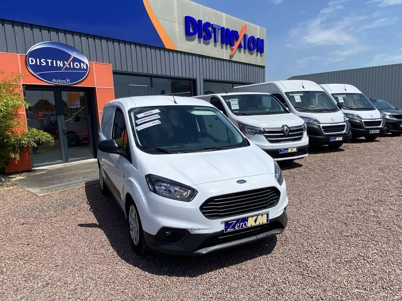 Ford Tourneo Courier blanc vue 3/4 avant droit, fourgon utilitaire devant un garage avec plusieurs véhicules blancs alignés.