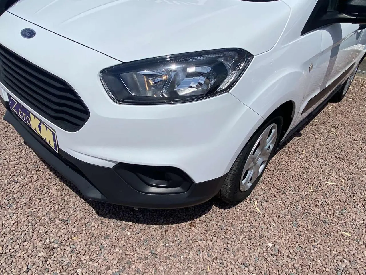 Vue 3/4 avant droit d'un Ford Tourneo Courier blanc avec enjoliveurs cache moyeu sur un sol gravillonné.