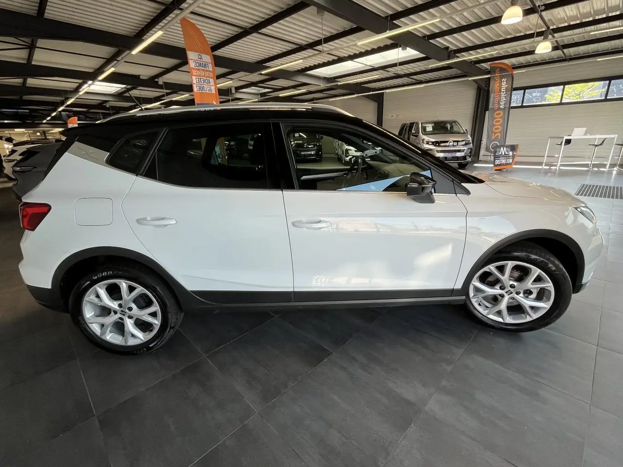 Vue latérale côté gauche d'un SEAT Arona blanc Nevada 2024 avec toit noir et jantes alliage 16 pouces en showroom.