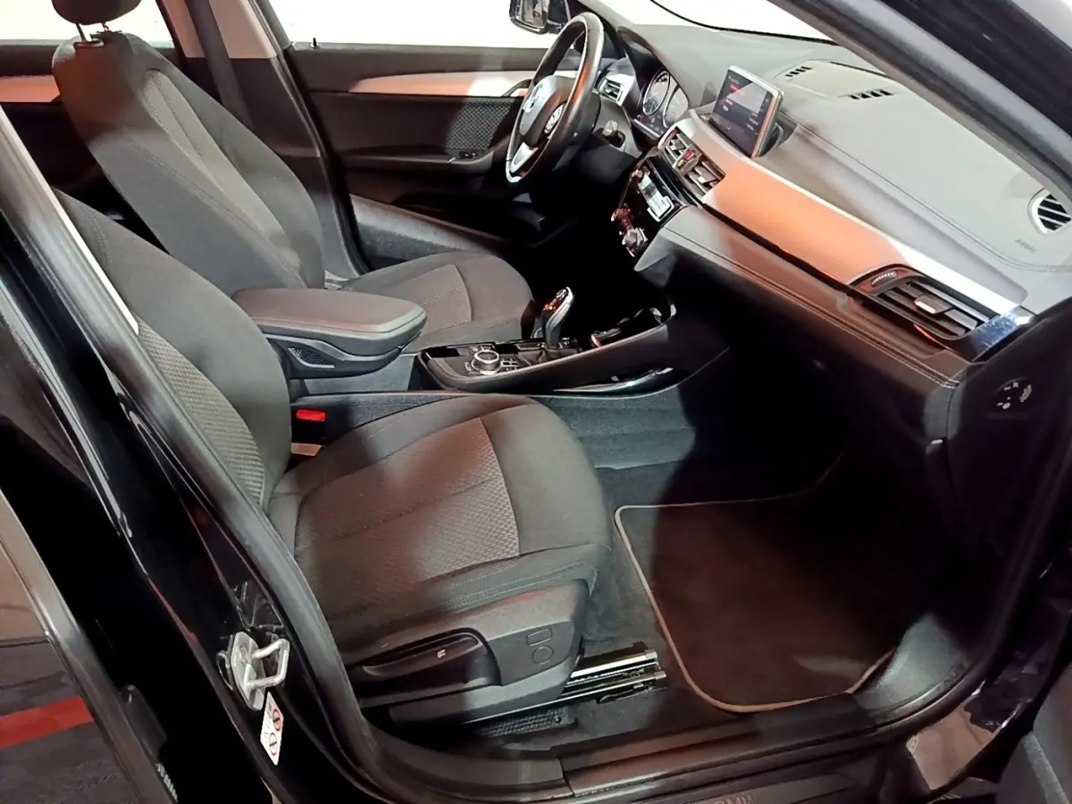 Vue intérieure côté conducteur de la BMW X2 sDrive18i Lounge noire, avec tableau de bord et sièges en tissu gris.