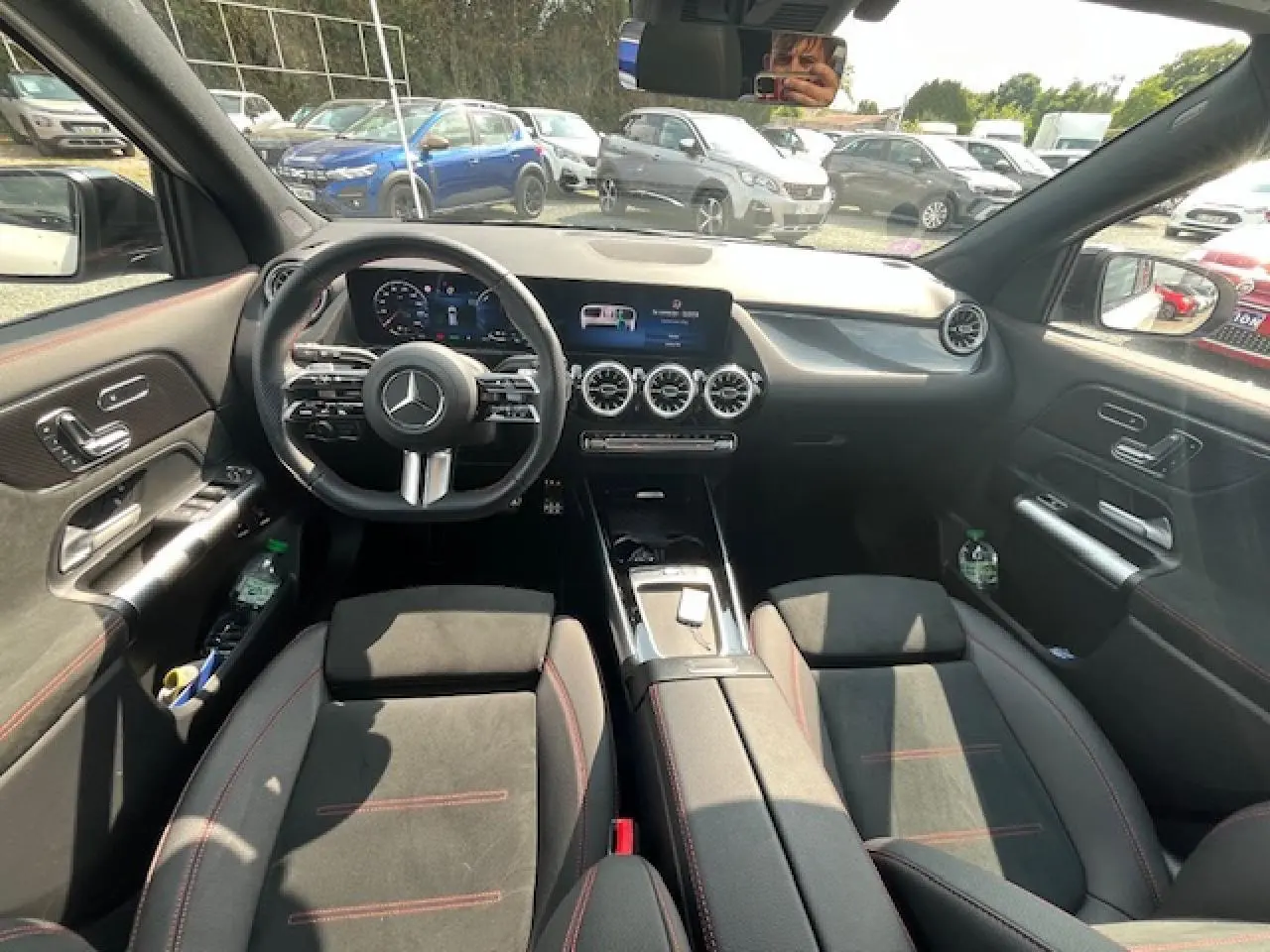 Intérieur du Mercedes GLA 250 e hybride 2024, vue avant du poste de conduite avec écran central et sièges noirs à surpiqûres rouges.