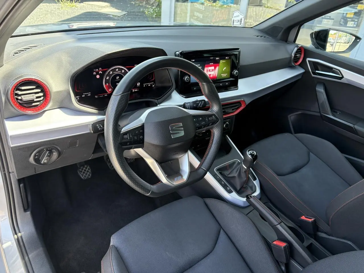 Intérieur de la SEAT Arona 2024 vu côté conducteur, volant sport cuir noir avec surpiqûres rouges et tableau de bord digital.