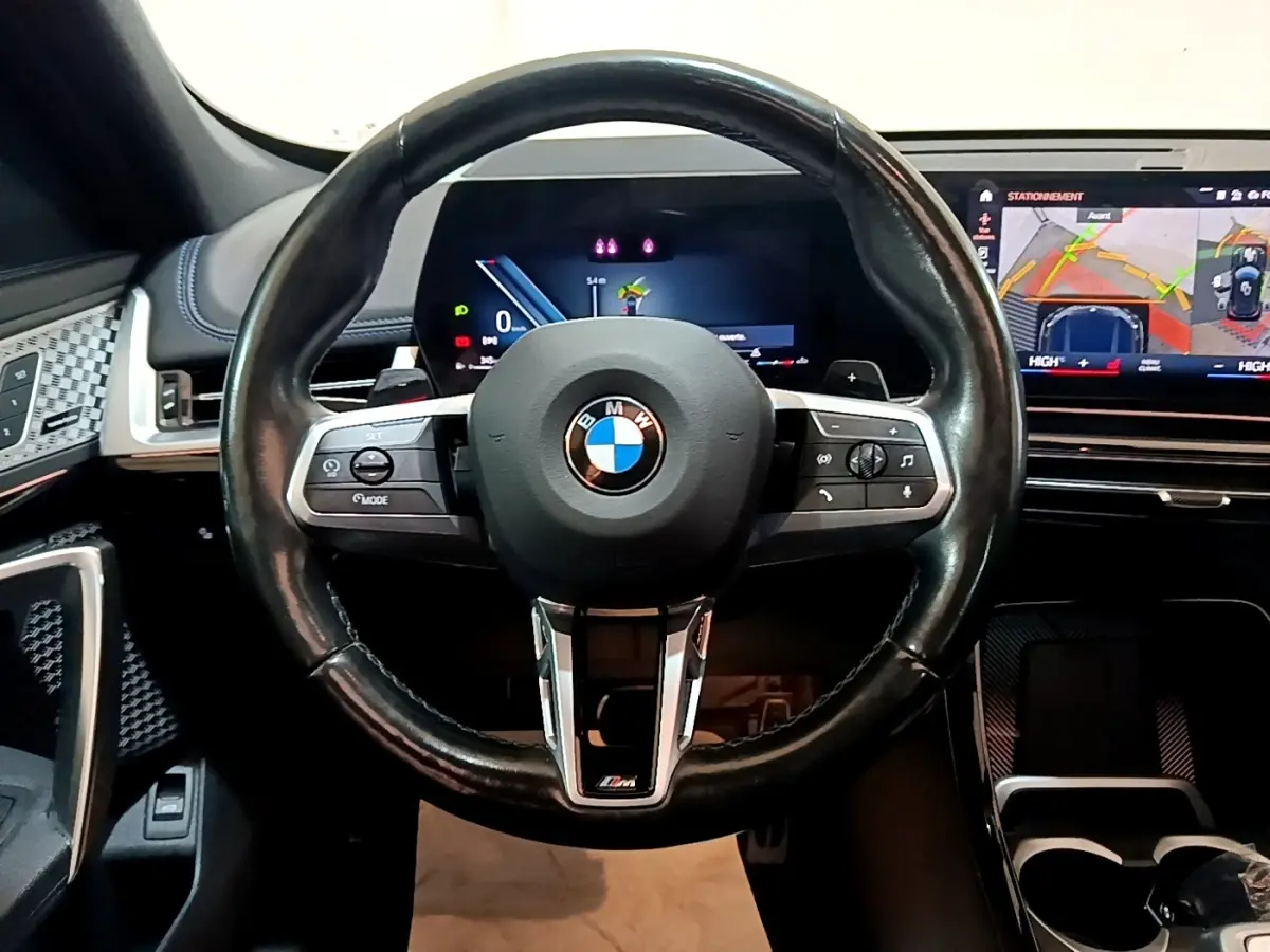 Vue intérieure centrée sur le volant cuir noir M Sport de la BMW X1 2023 avec tableau de bord digital et écran tactile.