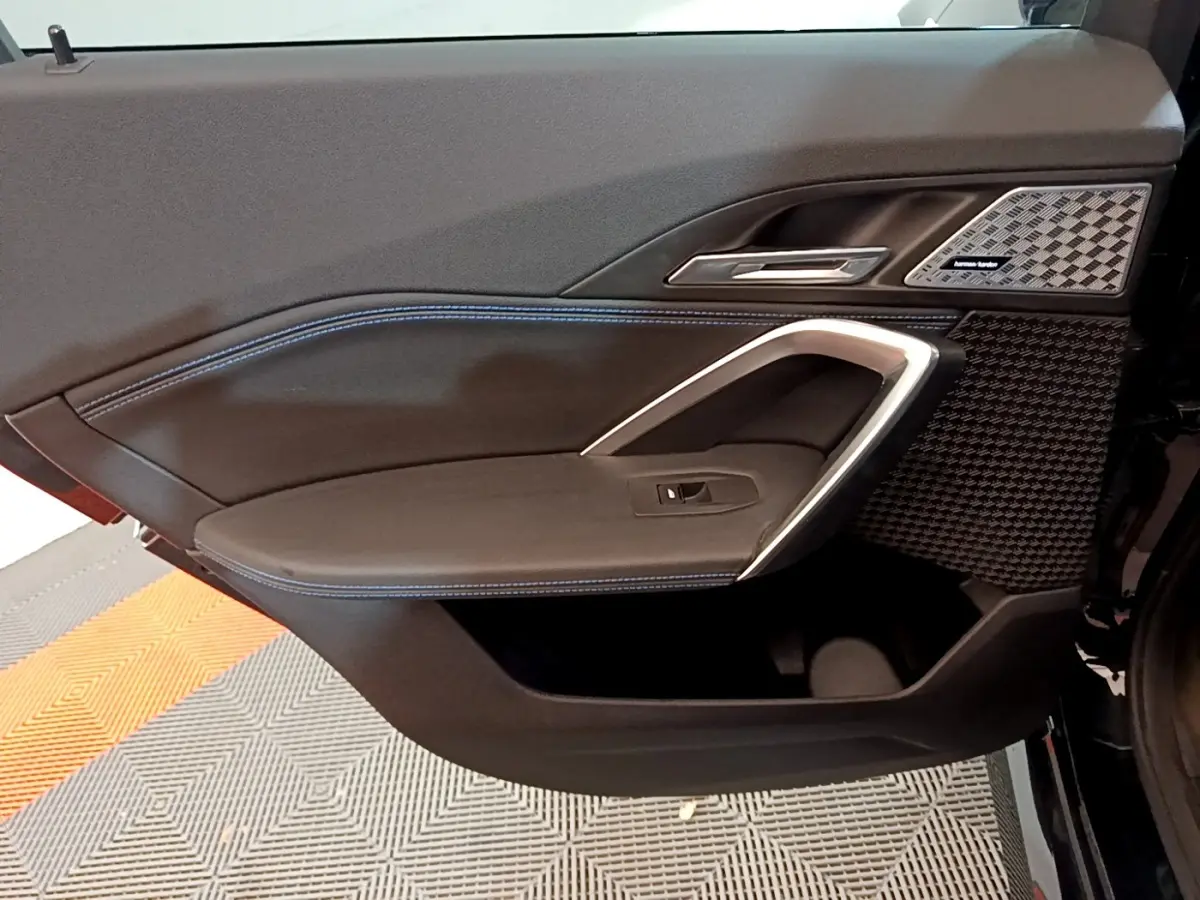 Intérieur côté gauche de porte BMW X1 noir, avec surpiqûres bleues et haut-parleur Harman Kardon visible.