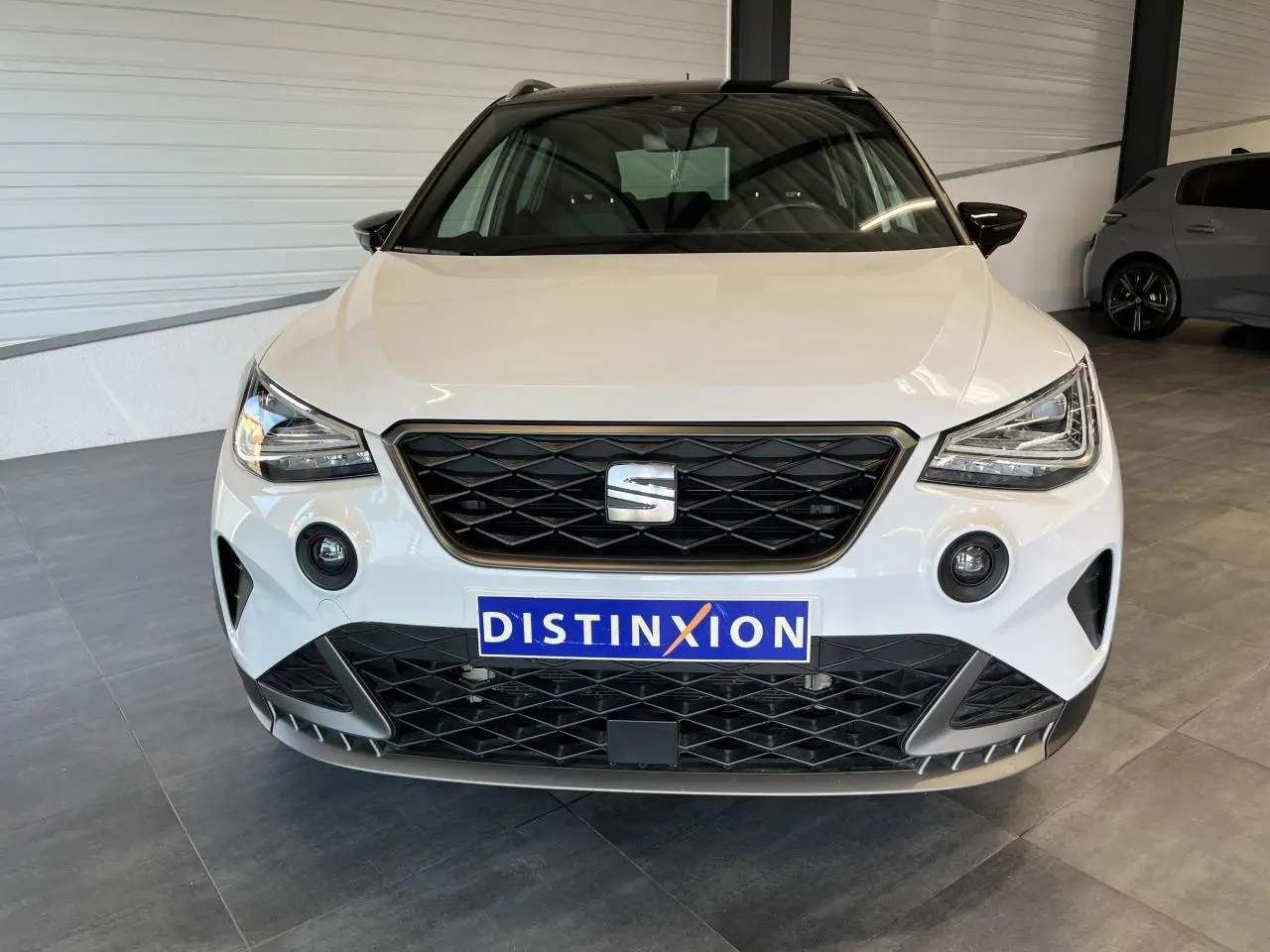 Vue frontale d'un SEAT Arona blanc Nevada avec calandre noire et logo central dans un showroom.
