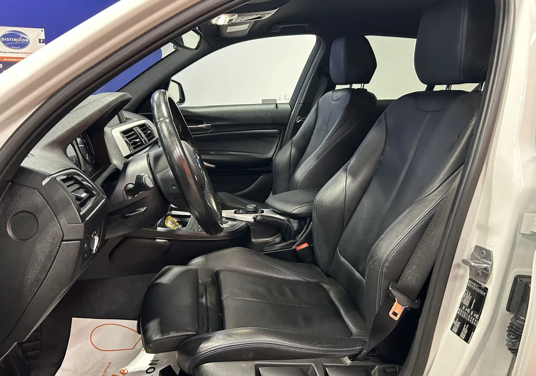 Vue intérieure côté conducteur d'une BMW Série 1 blanche de 2019, sièges en cuir noir avec coutures bleues et volant multifonction.