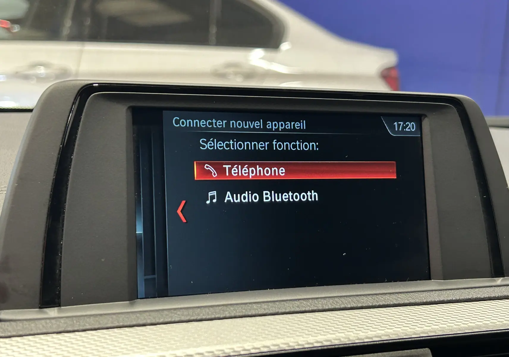 Écran central de la BMW Série 1 blanc 120iA M Sport affichant le menu Bluetooth avec une voiture blanche en arrière-plan.