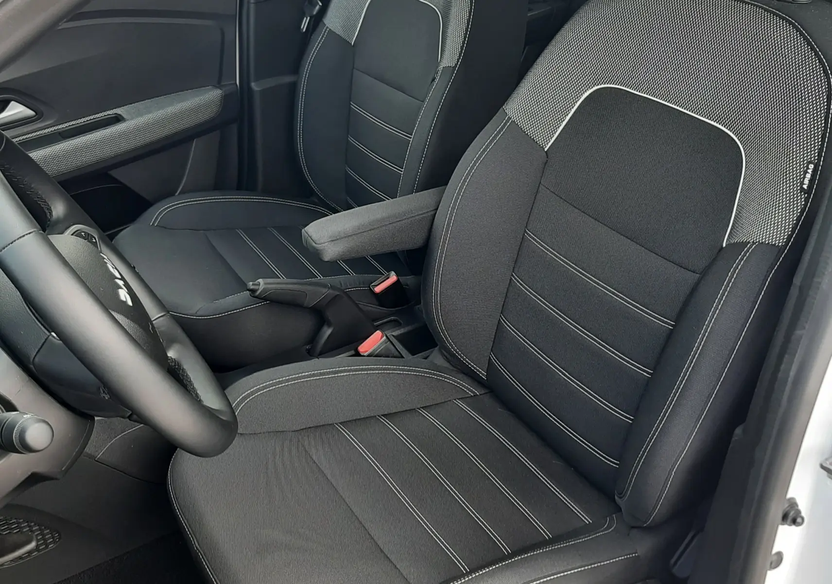 Vue intérieure côté conducteur de la Dacia Sandero 2024, sièges tissu noir avec surpiqûres blanches et accoudoir central.