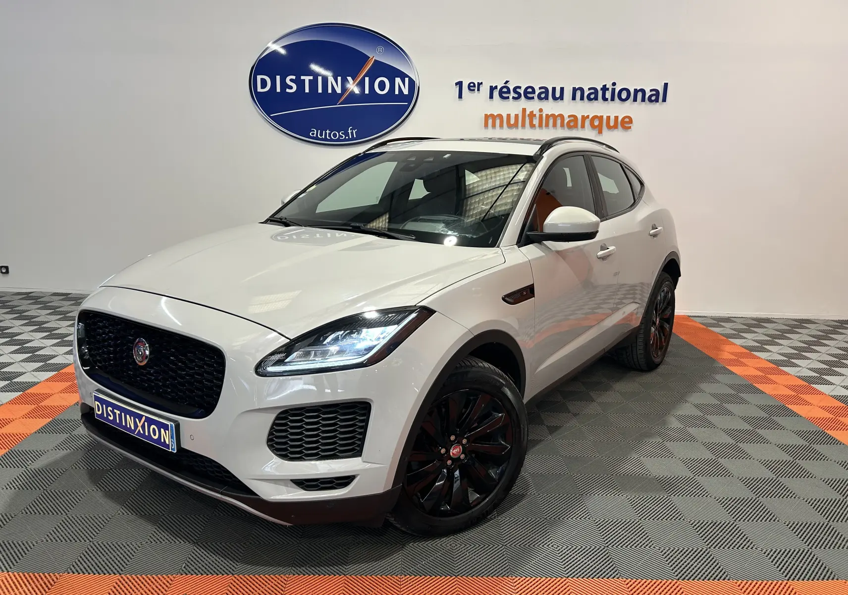 Jaguar E-PACE beige vue 3/4 avant droit avec jantes noires et toit panoramique en intérieur showroom.
