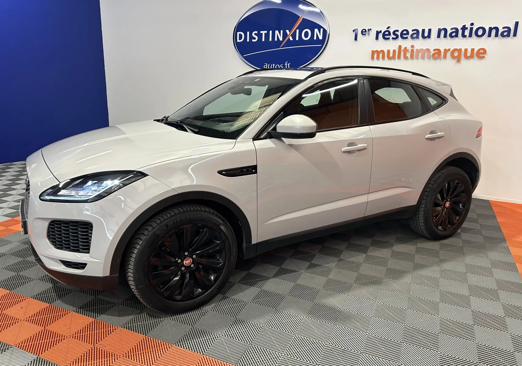 Jaguar E-Pace beige vue 3/4 avant droit, jantes noires brillantes, toit panoramique et hayon électrique visibles.