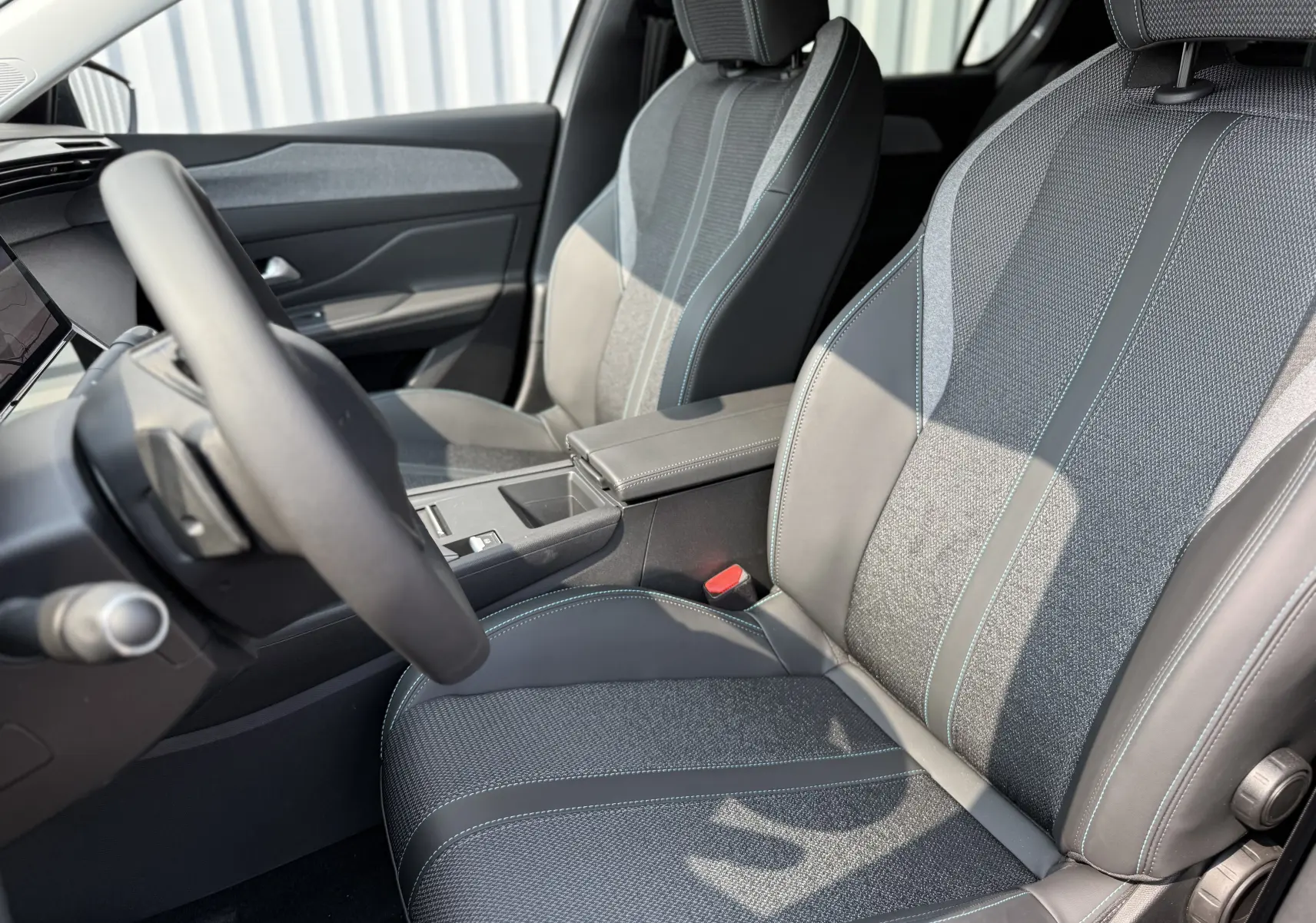 Intérieur de la Peugeot 308 2025, vue côté conducteur, sièges en tissu gris avec surpiqûres bleues et cuir noir.
