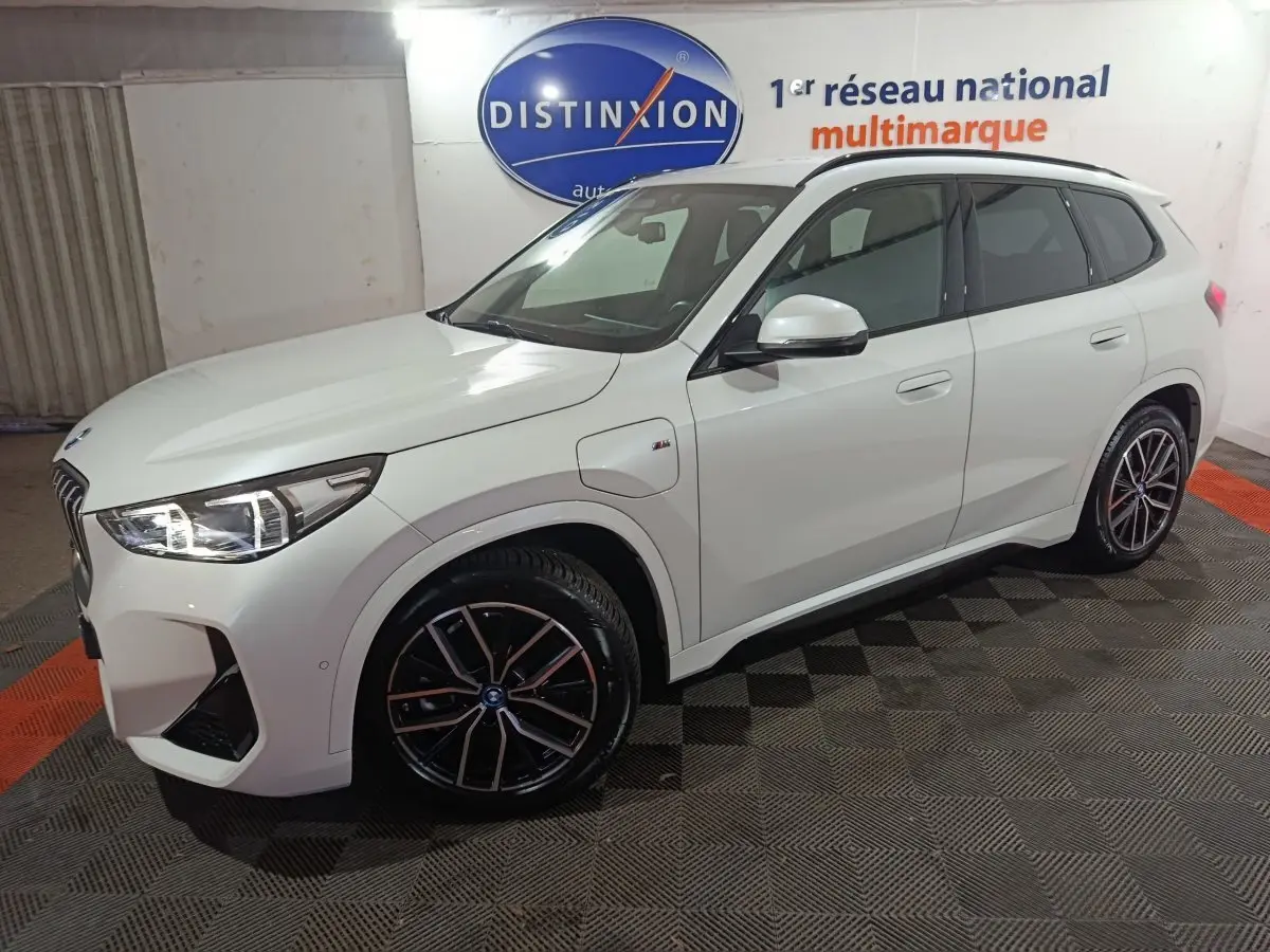 BMW X1 xDrive25e blanc en 3/4 avant droit, avec jantes noires et éclairage LED allumé.