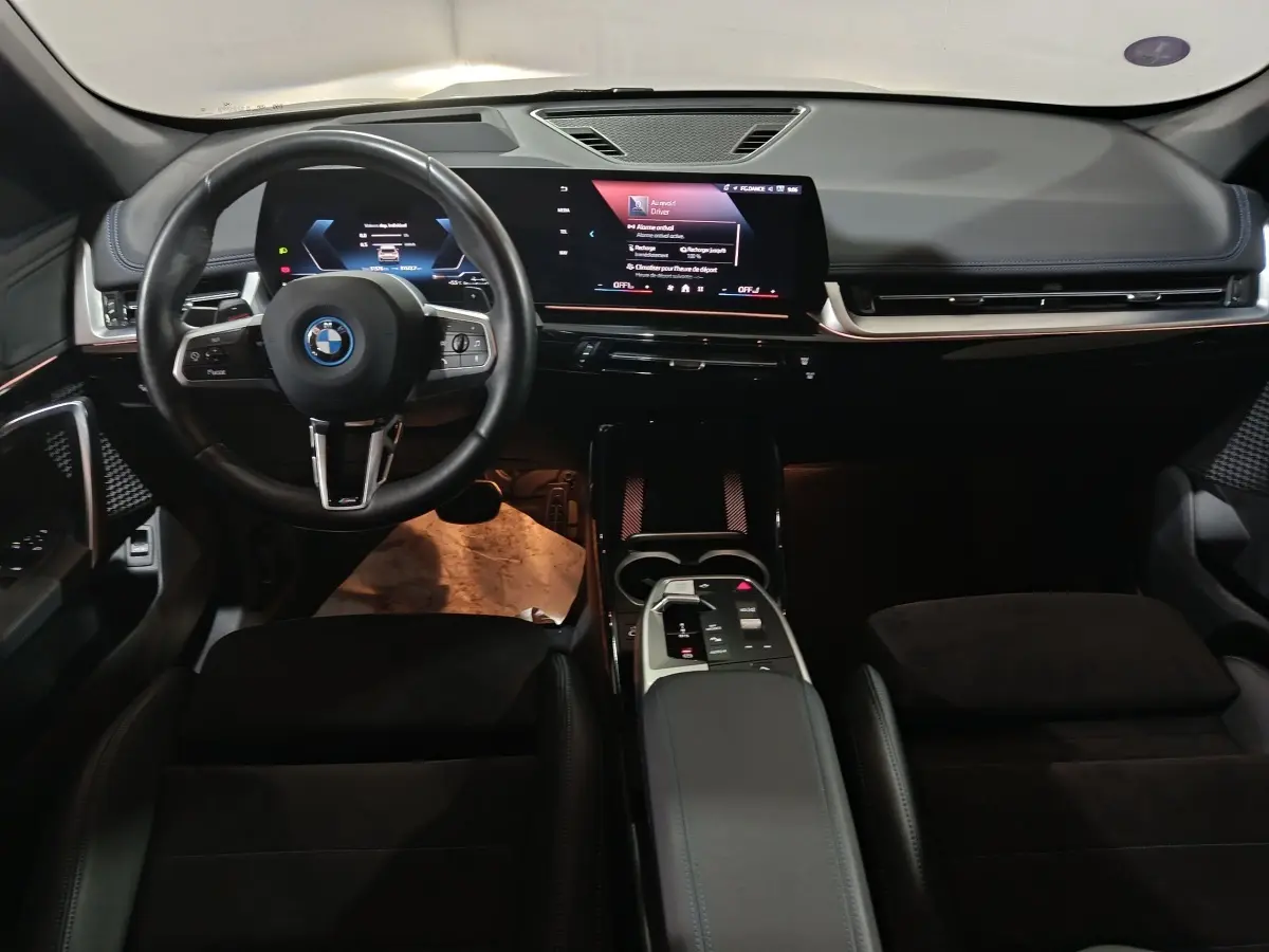 Vue intérieure avant du BMW X1 xDrive25e M Sport 2024, tableau de bord digital et volant cuir noir avec logo BMW.