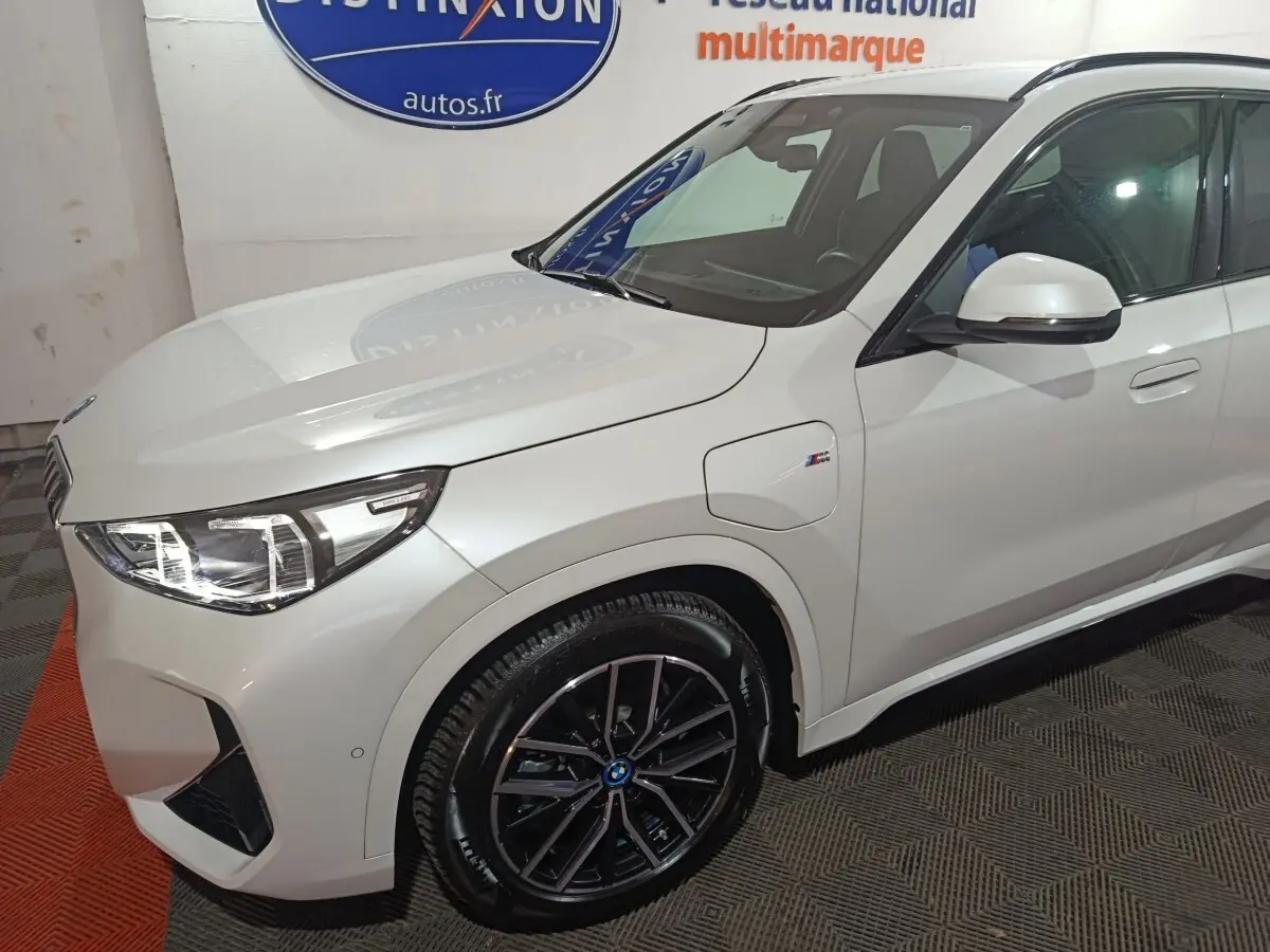Vue 3/4 avant droit d’un BMW X1 blanc hybride rechargeable avec jantes noires et badge M Sport visible sur l’aile.