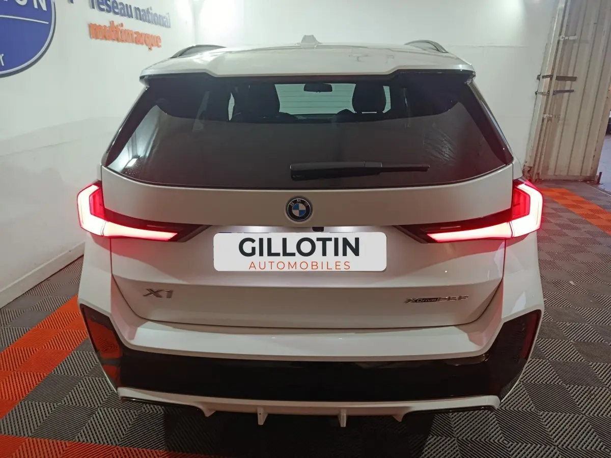 Vue arrière d’un BMW X1 blanc 2024 avec feux LED allumés et logo xDrive25e M Sport visible.