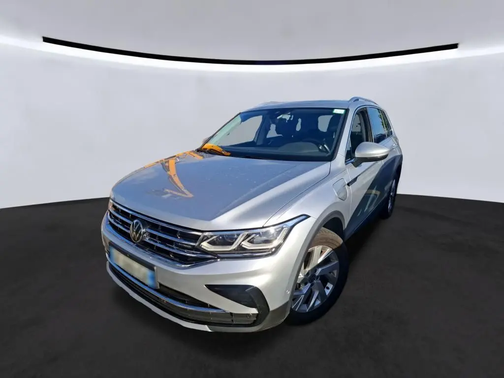 Vue 3/4 avant d'un Volkswagen Tiguan gris clair hybride rechargeable, avec calandre chromée et phares LED visibles.