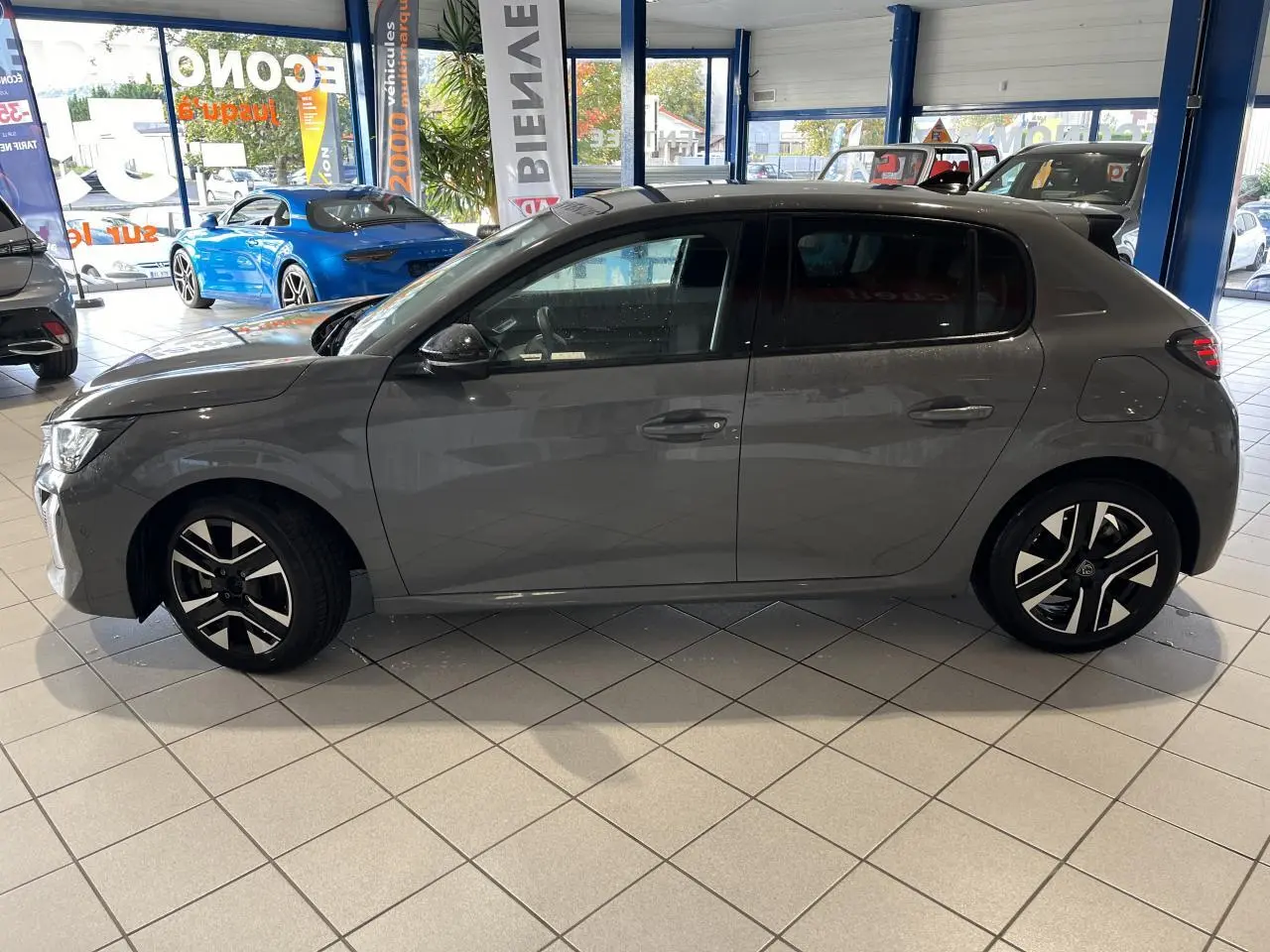 Profil droit d'une Peugeot 208 1.2 essence gris clair Selenium avec jantes alliage bi-tons, en intérieur showroom.