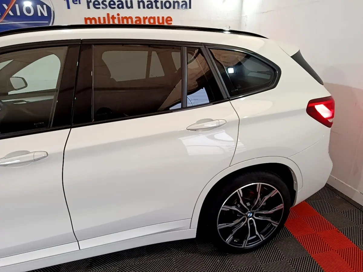 Vue de côté droit arrière d’un BMW X1 blanc avec jantes alliage noires et feux arrière allumés.