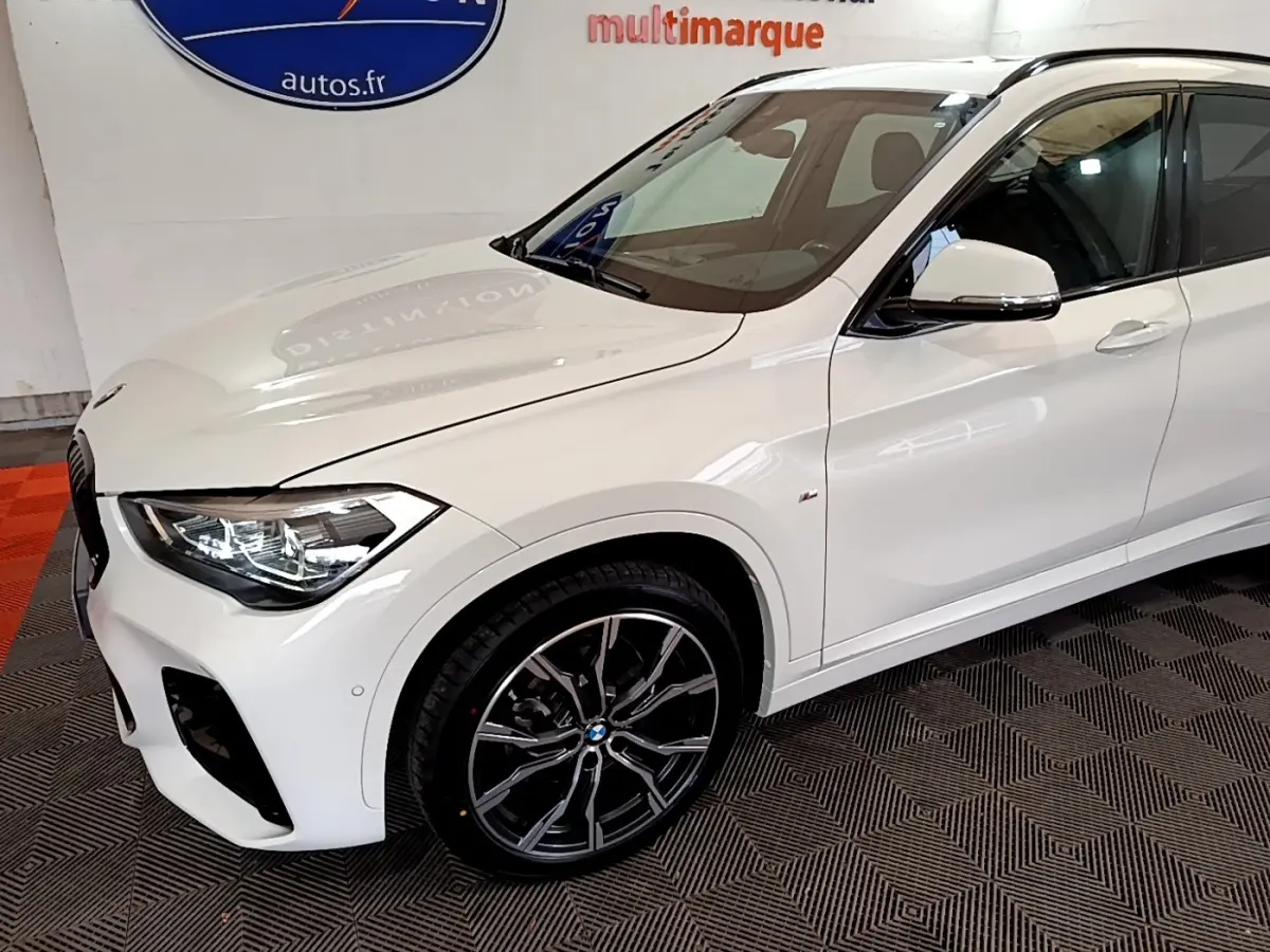 Vue 3/4 avant gauche d’un BMW X1 blanc sDrive18i M Sport avec jantes alliage noires et phares LED allumés.