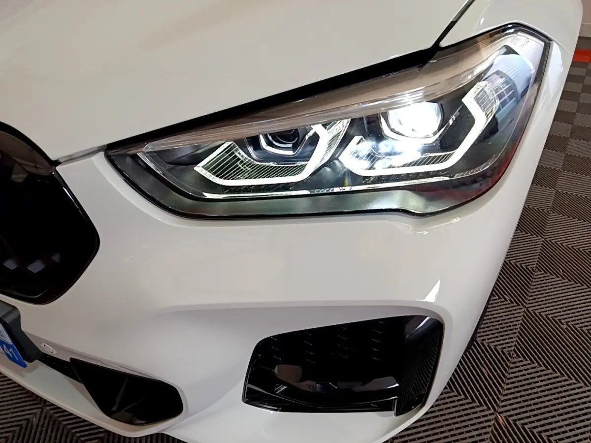 Gros plan sur le phare avant droit allumé d’un BMW X1 sDrive18i M Sport blanc, avec calandre noire partielle visible.