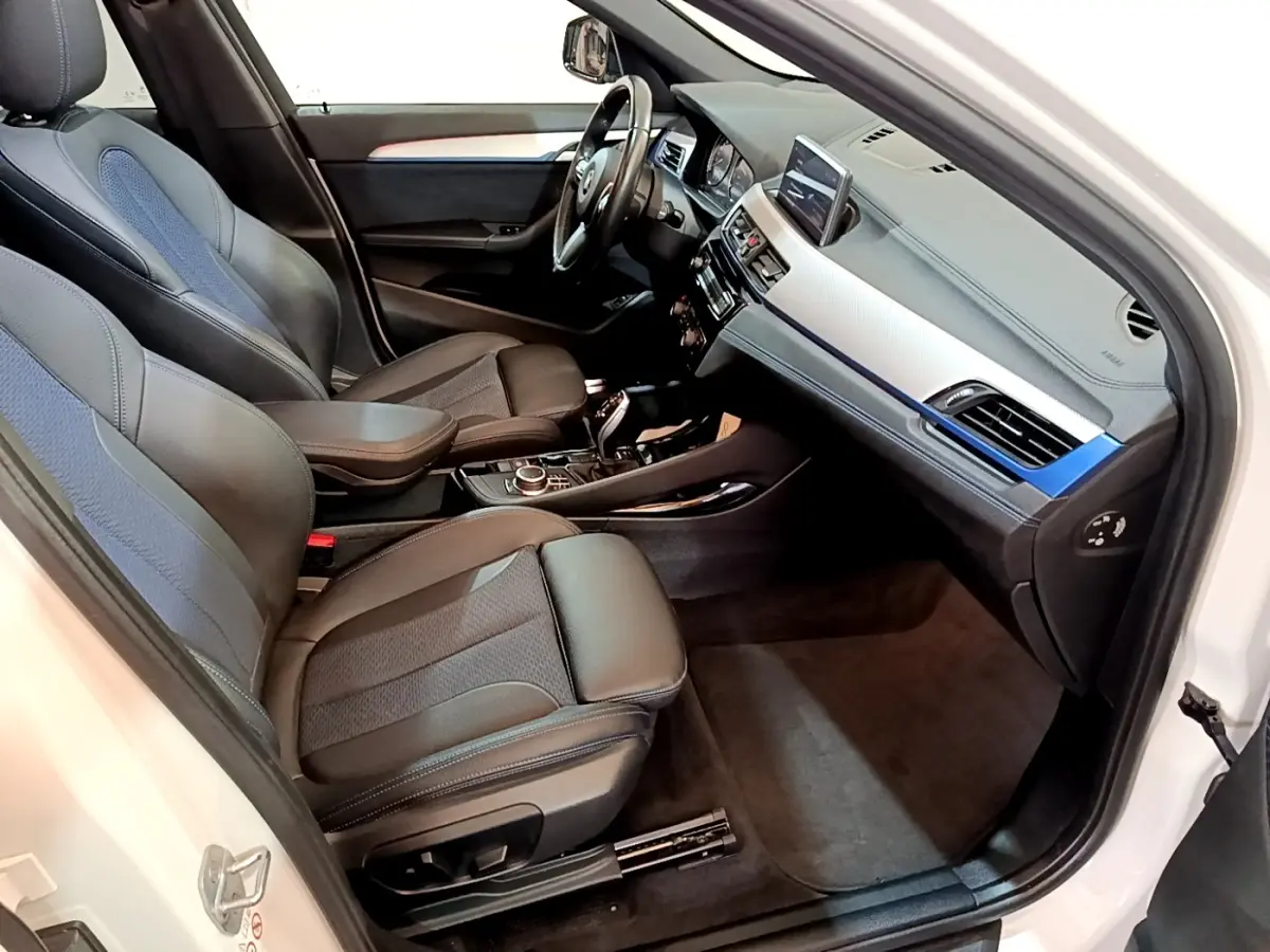 Vue intérieure côté conducteur d’un BMW X1 blanc 2022, sièges cuir noir avec inserts tissu bleu et tableau de bord moderne.