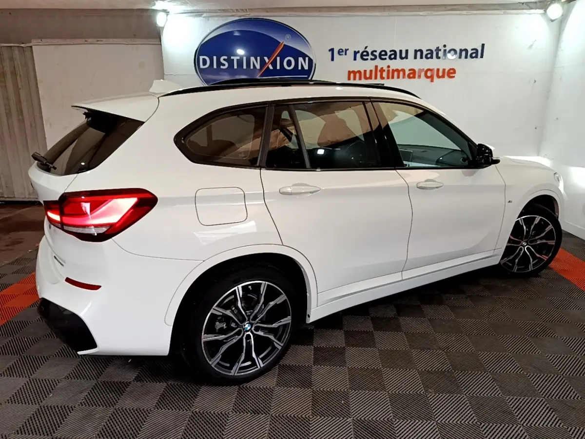 Vue 3/4 arrière droite d’un BMW X1 blanc sDrive18i M Sport avec jantes noires et feux arrière allumés.