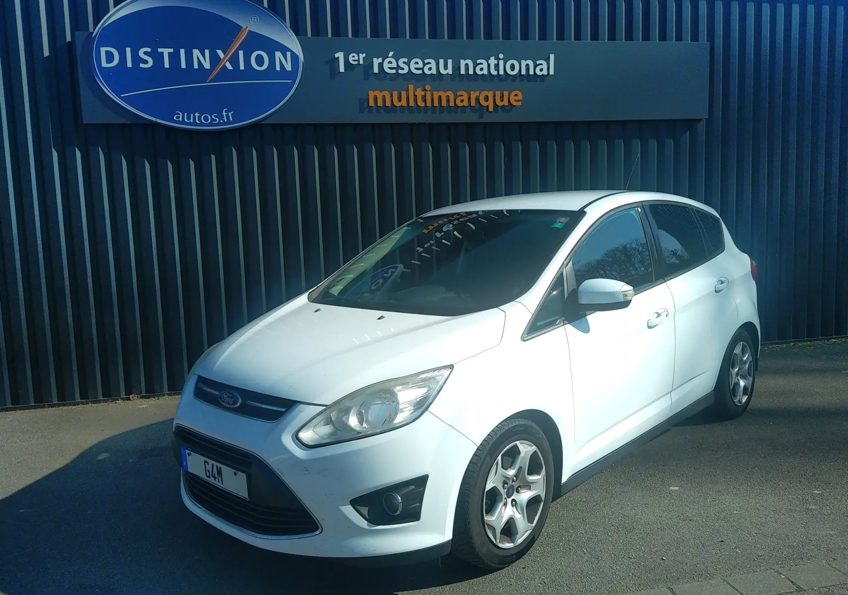 Ford C-MAX blanc vue 3/4 avant droit, garé devant un mur noir avec enseigne Distinxion.