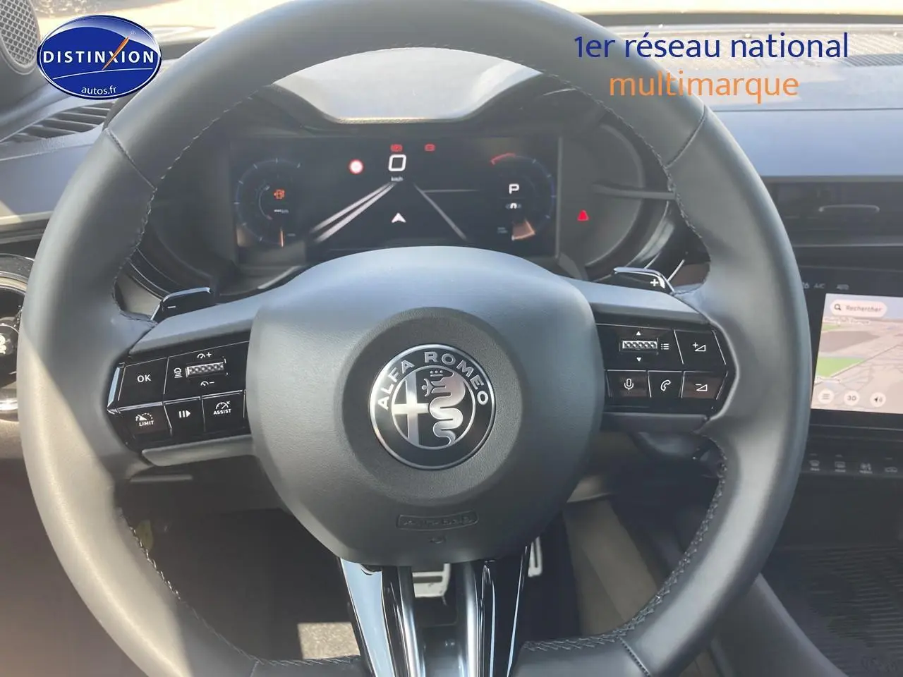 Vue rapprochée du volant noir de l'Alfa Romeo Junior 1.2 hybride 2026 avec tableau de bord digital et écran tactile visible.