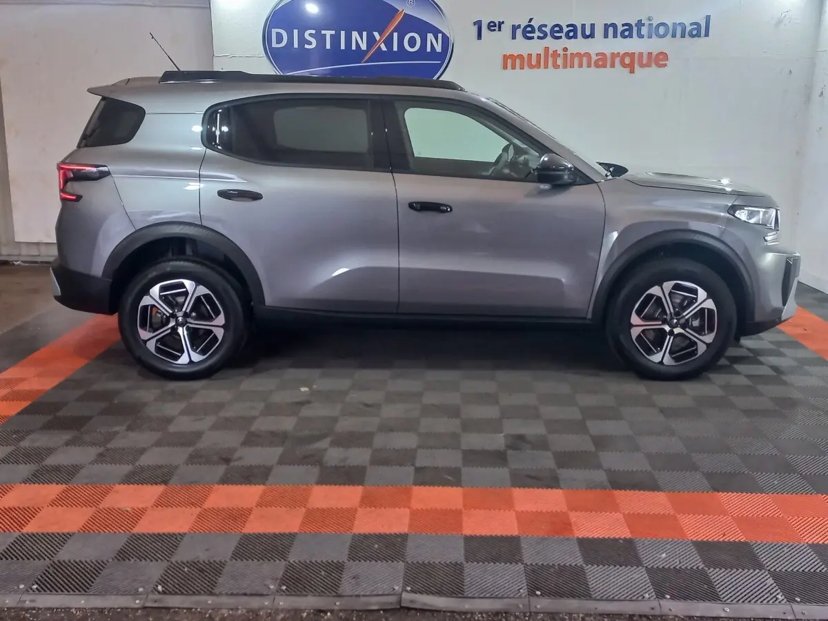 Vue latérale droite d'un Citroën C3 hybride gris foncé, stationné en intérieur sur sol damier noir et orange.