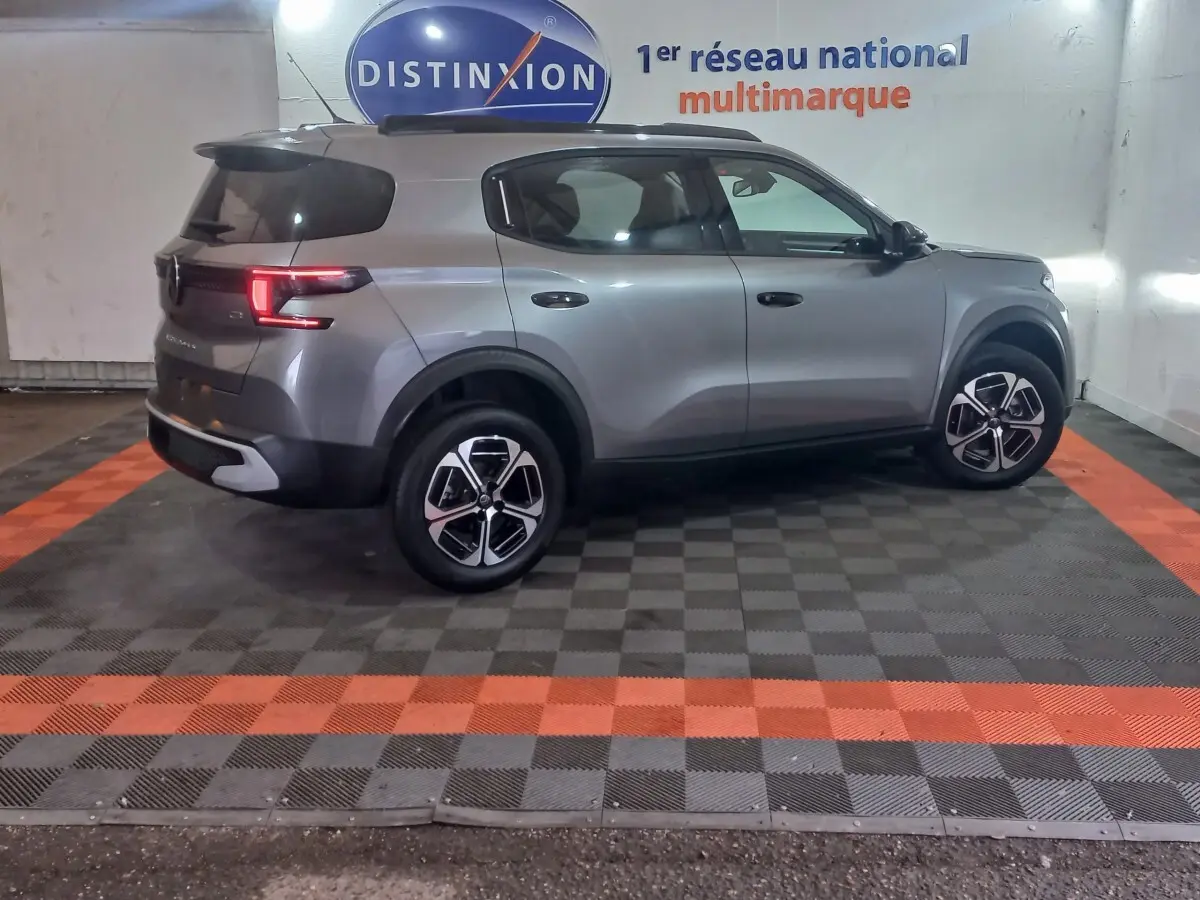 Vue latérale droite d'une Citroën C3 hybride gris foncé, stationnée en intérieur sur un sol à damiers noir et orange.