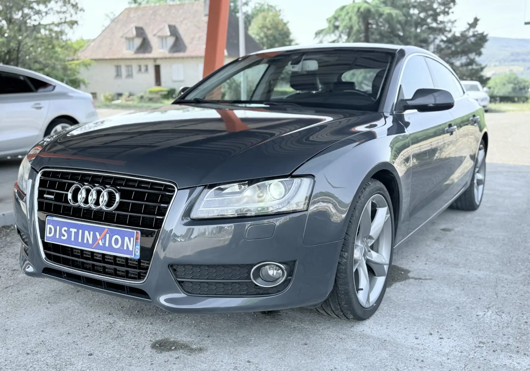 Audi A5 Sportback gris dauphin vue 3/4 avant droit, avec calandre noire et phares allumés sous abri.
