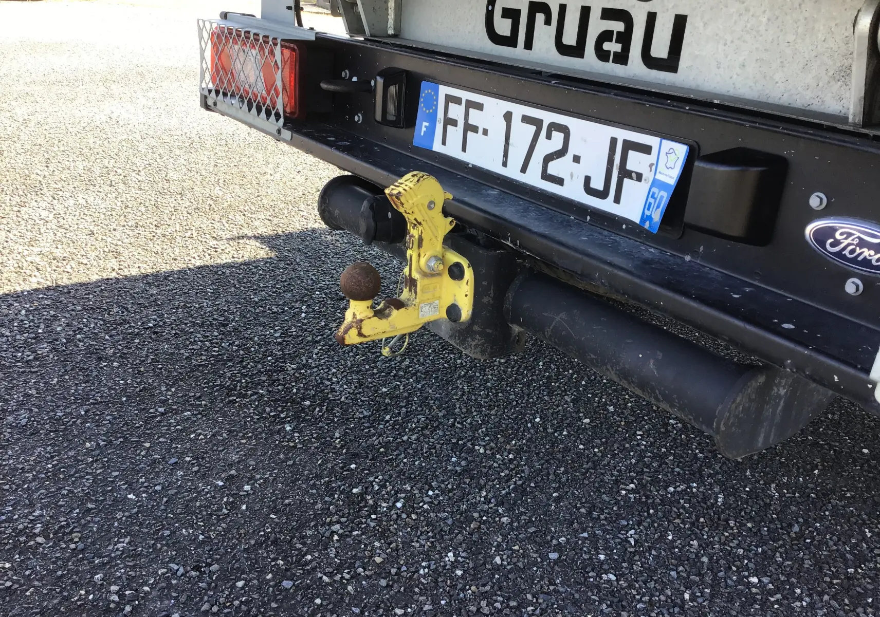 Gros plan sur l'attelage arrière jaune rouillé d'un Ford Transit blanc avec plaque d'immatriculation française visible.
