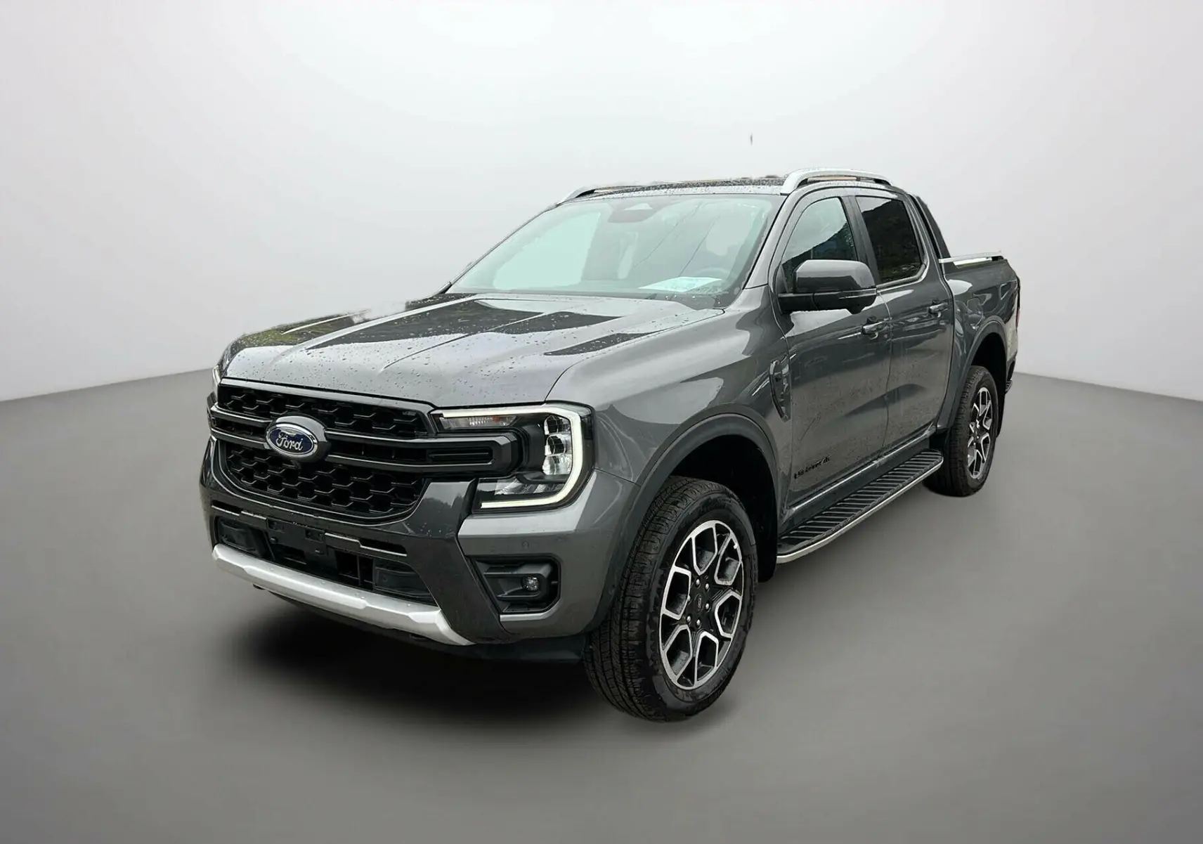 Ford Ranger 3.0 V6 240 Wildtrak Plus gris carbone vu en 3/4 avant droit avec jantes alliage et barres de toit visibles.
