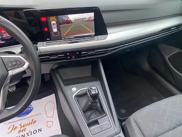 Vue intérieure du tableau de bord et console centrale d'une Volkswagen Golf VIII gris argent, avec levier de vitesse manuel et écran multimédia affichant la caméra de recul.