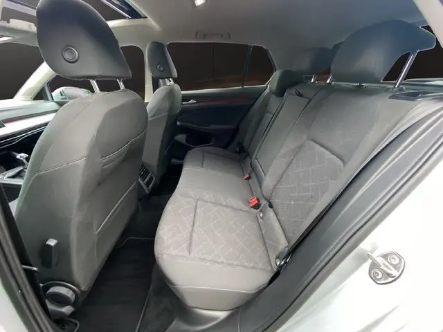 Vue intérieure arrière droite de la Volkswagen Golf VIII gris argent, sièges tissu noir et toit panoramique ouvrant visible.