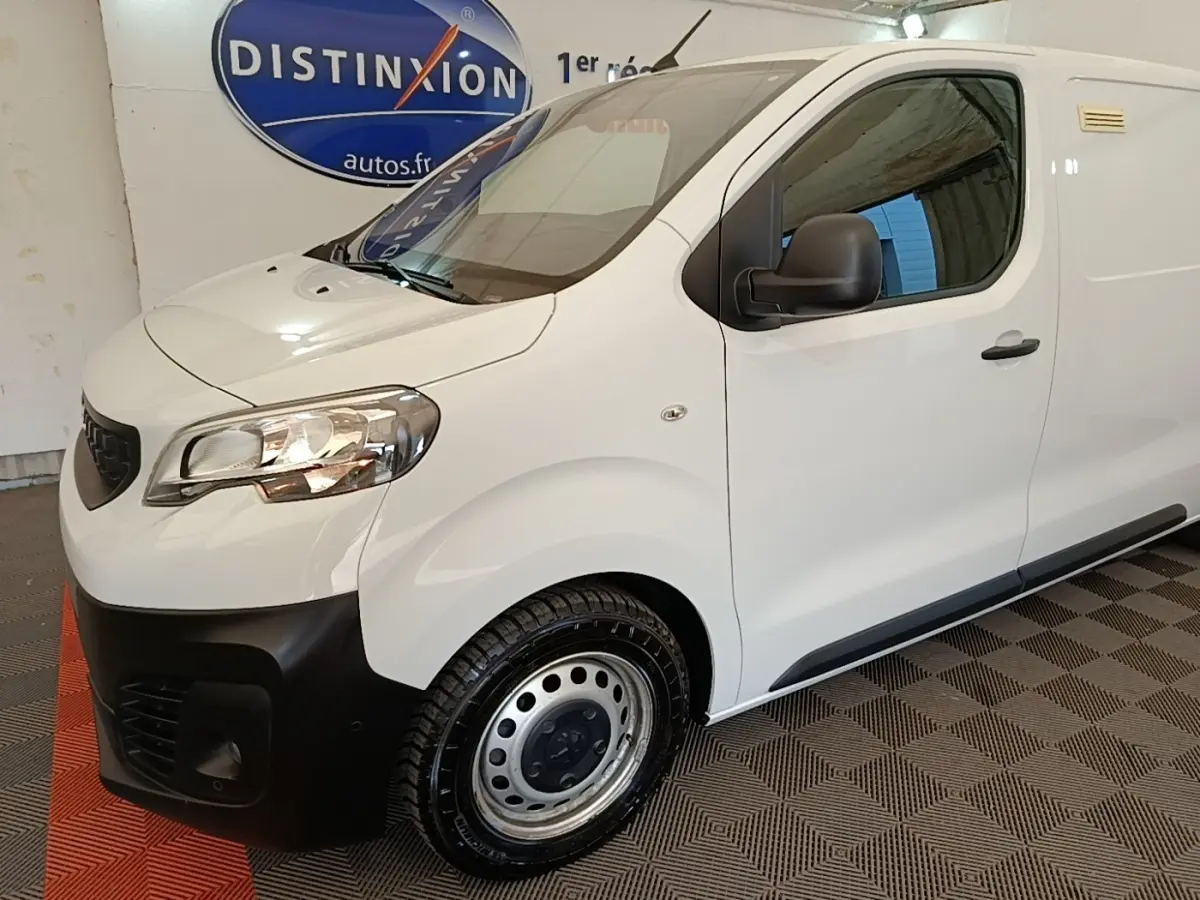 Vue 3/4 avant droit d’un Peugeot Expert Fourgon blanc avec pare-chocs noir et jantes acier en intérieur showroom.