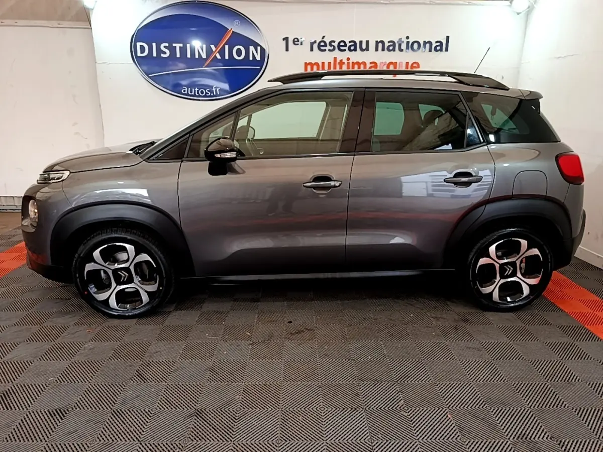 Vue de profil côté gauche d’un Citroën C3 Aircross gris foncé avec barres de toit noires en intérieur showroom.