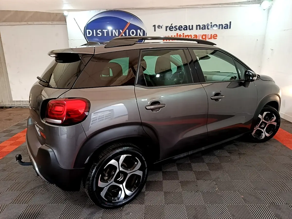 Vue 3/4 arrière droit d'un Citroën C3 Aircross gris foncé avec jantes bicolores et barres de toit noires.