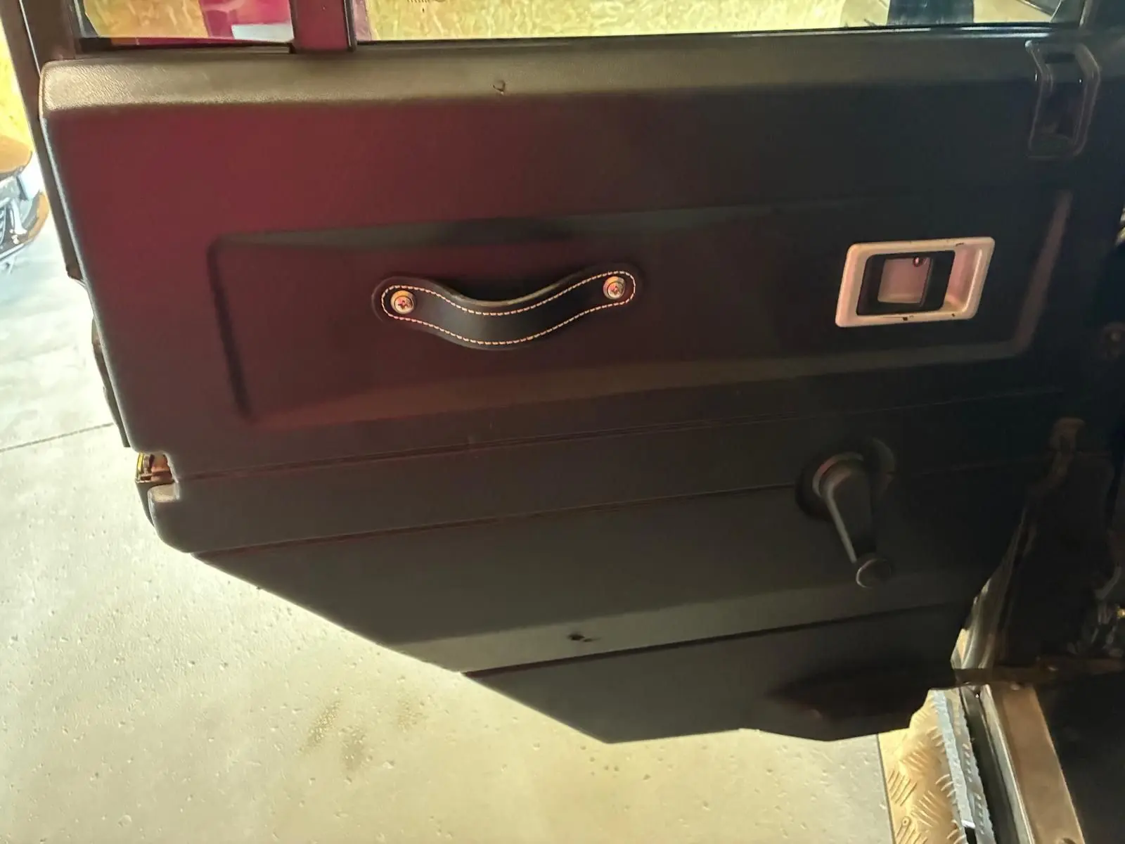 Détail de la porte intérieure côté gauche du Land Rover Defender 110 vert de 2005 avec poignée en cuir et manivelle de vitre.