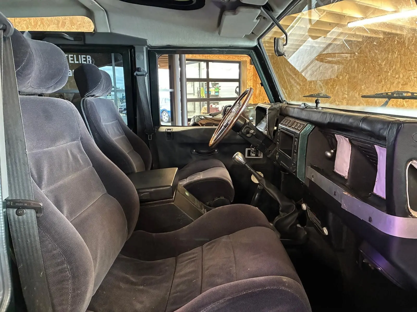 Vue intérieure côté conducteur du Land Rover Defender 110 2005, montrant les sièges noirs et le tableau de bord avec volant bois.
