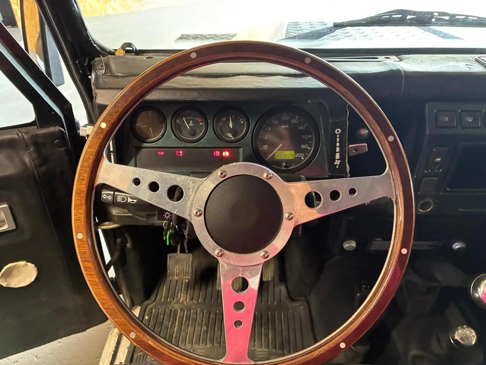 Vue intérieure centrée sur le volant bois du Land Rover Defender 110 vert de 2005 avec tableau de bord et pédales visibles.