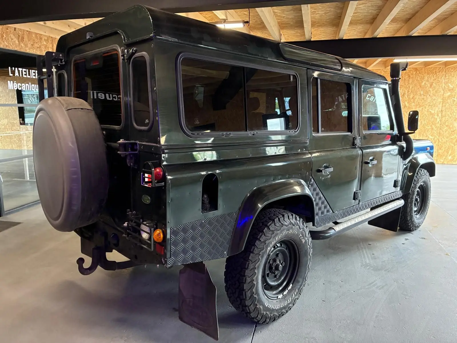 Land Rover Defender 110 vert vu en 3/4 arrière droit, avec roue de secours et protections en métal sur les bas de caisse.